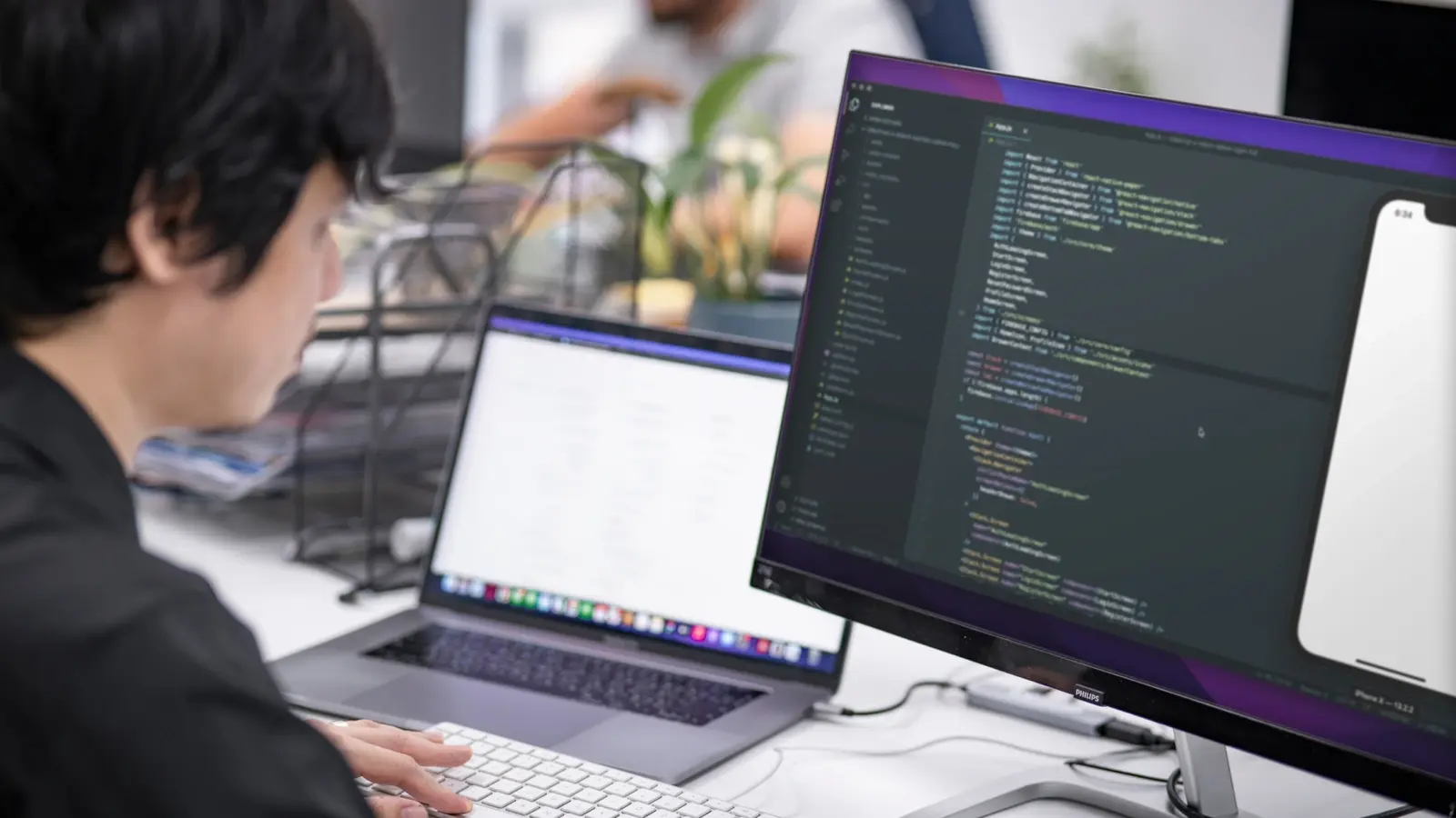 A software developer builds a mobile application with code and a smartphone preview displayed on a monitor.