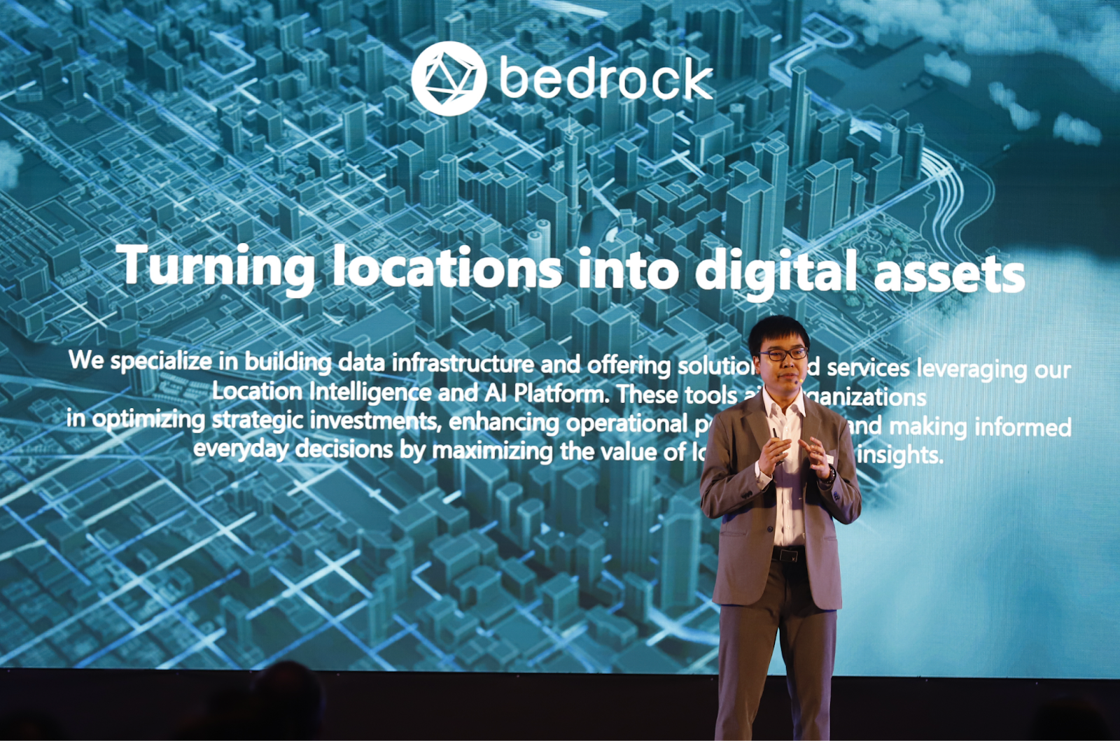 Dr. Chakaphan Chullabodhi, the Product Development Manager at Bedrock Analytics, is on stage presenting a slide at the HERE Technologies event in Bangkok. The slide's title is "Turning locations into digital assets" over a blue, digital city illustration.